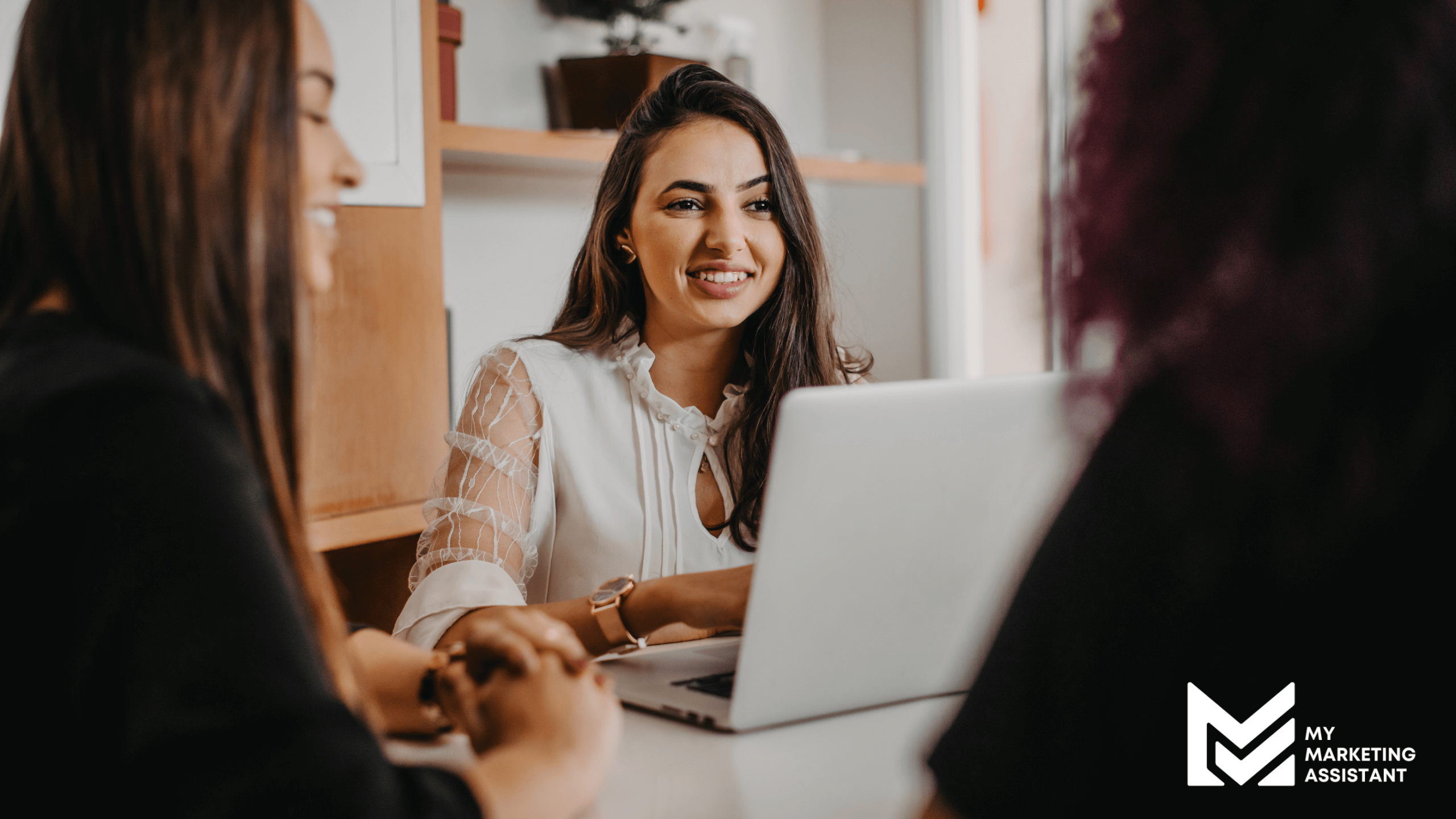A group of women seated around a table in an office, actively engaged in discussion with laptops open.