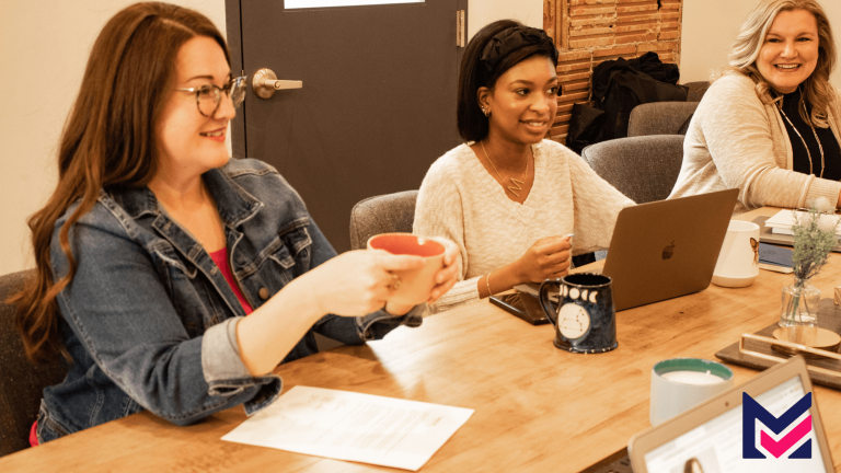 MYMA team gathered around a table with laptops, mugs, and notebooks, engaged in meeting discussion