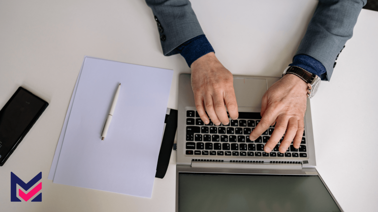 "Close-up of a person's hand using a laptop at a table, with paper, pen, and phone nearby.
