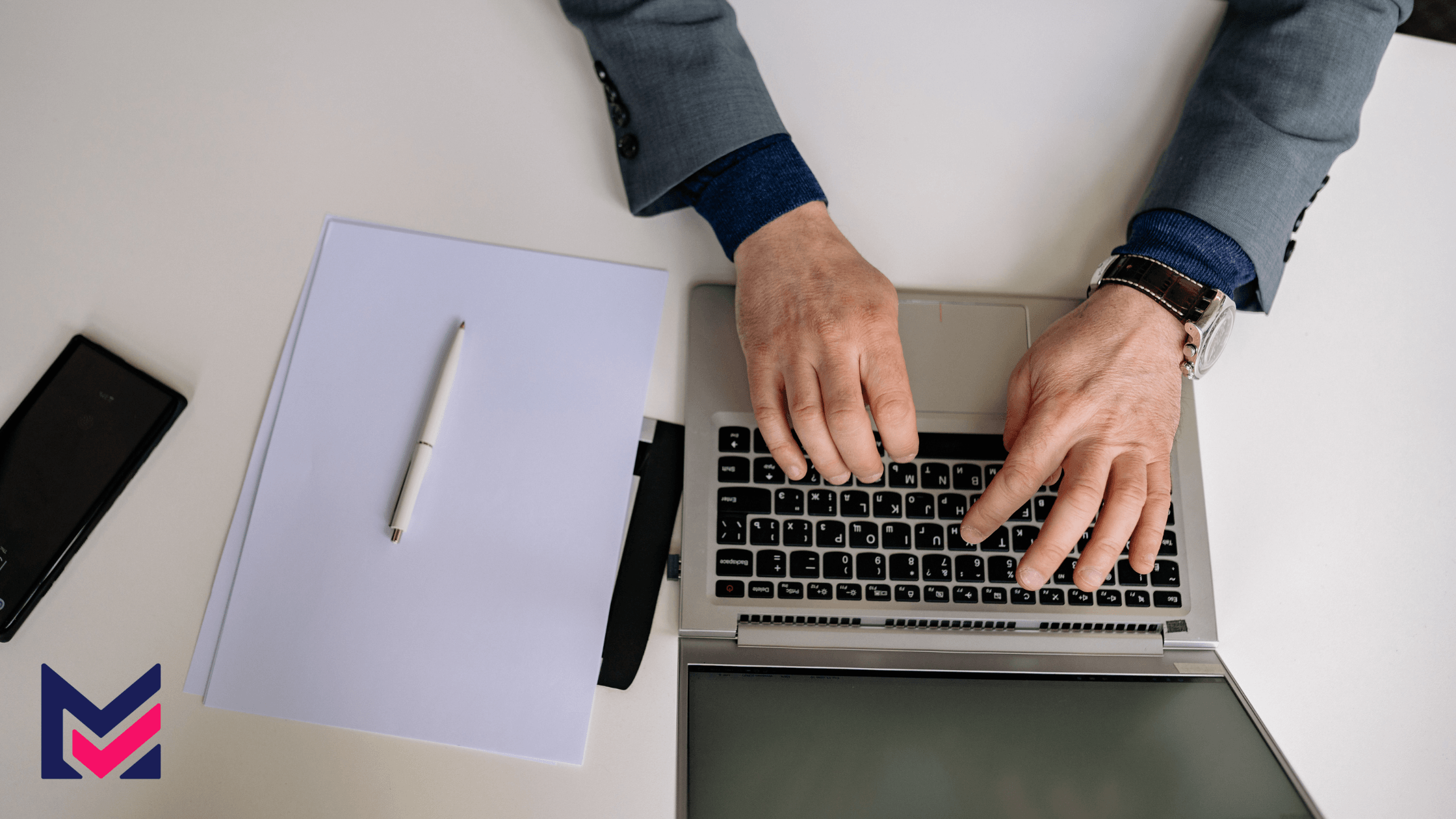 "Close-up of a person's hand using a laptop at a table, with paper, pen, and phone nearby.