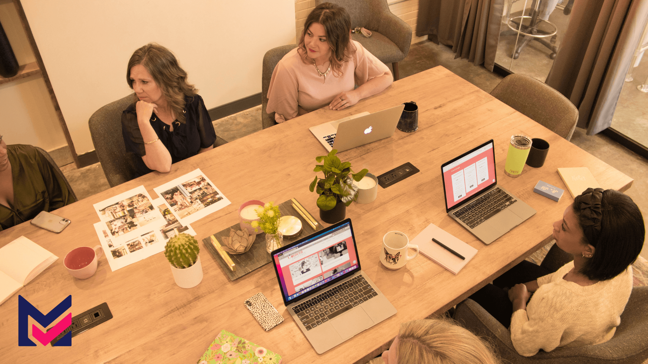 Team seated around a meeting table with laptops, mugs, ballpens, and notebooks.